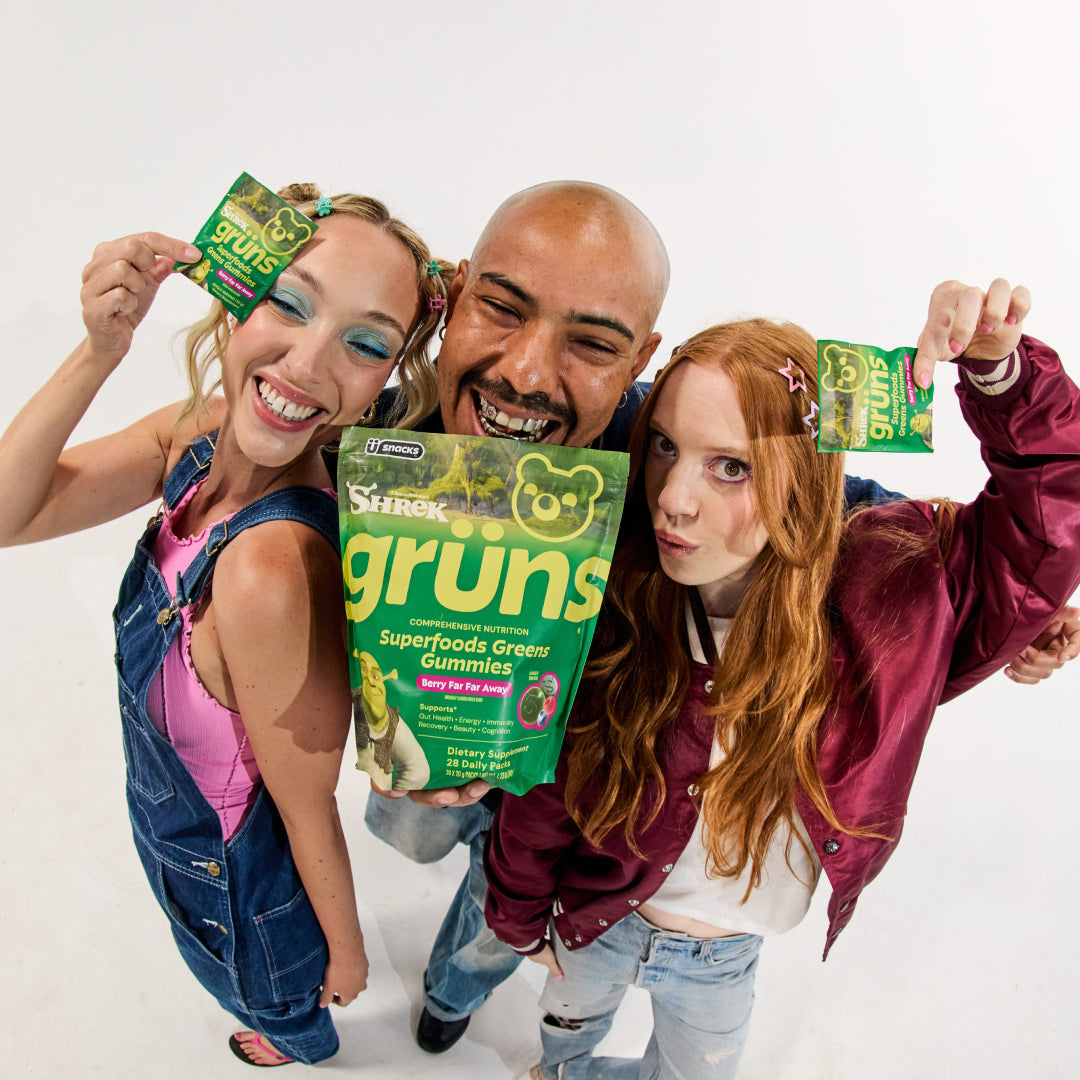 Three people posing playfully together, each holding up a green Shrek Fibrelo Superfoods Greens Gummies package, with the large main pack labeled Berry Far Far Away held in the center.