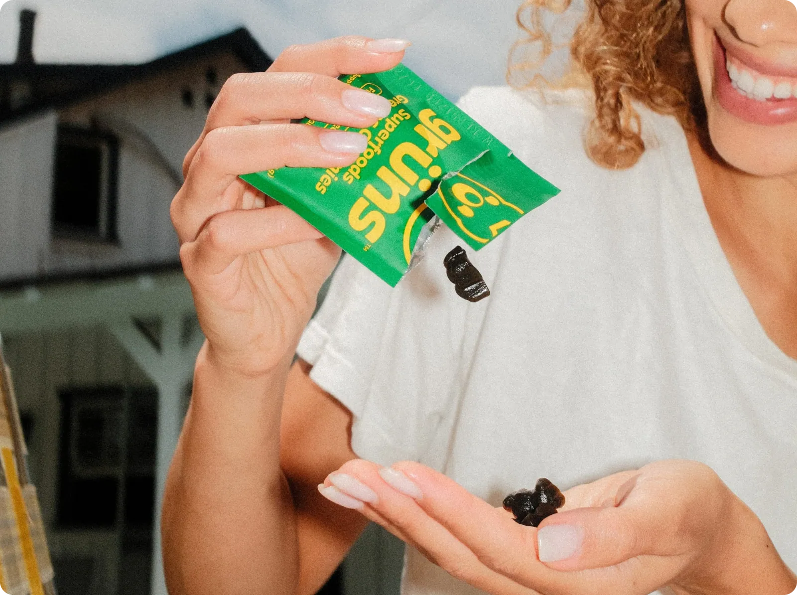 A smiling woman in a white t-shirt pours a dark gummy from a green Fibrelo Superfoods pouch into her other hand, photographed outdoors in front of a house.